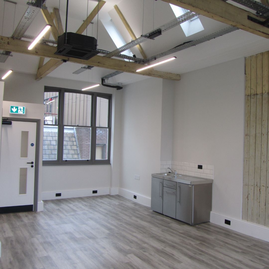 a bright and spacious modern office interior featuring exposed wooden beams, skylights, LED lighting, a compact kitchenette, and laminate flooring, offering a clean and functional workspace.